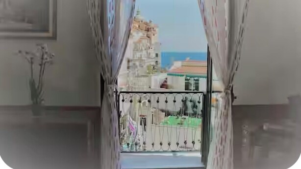 A view through a window with curtains, revealing a balcony with a metal railing and a building with a green roof in the background.