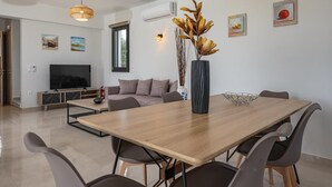 A modern living room with a wooden dining table, grey chairs, a flat-screen TV, a sofa, and decorative wall art.