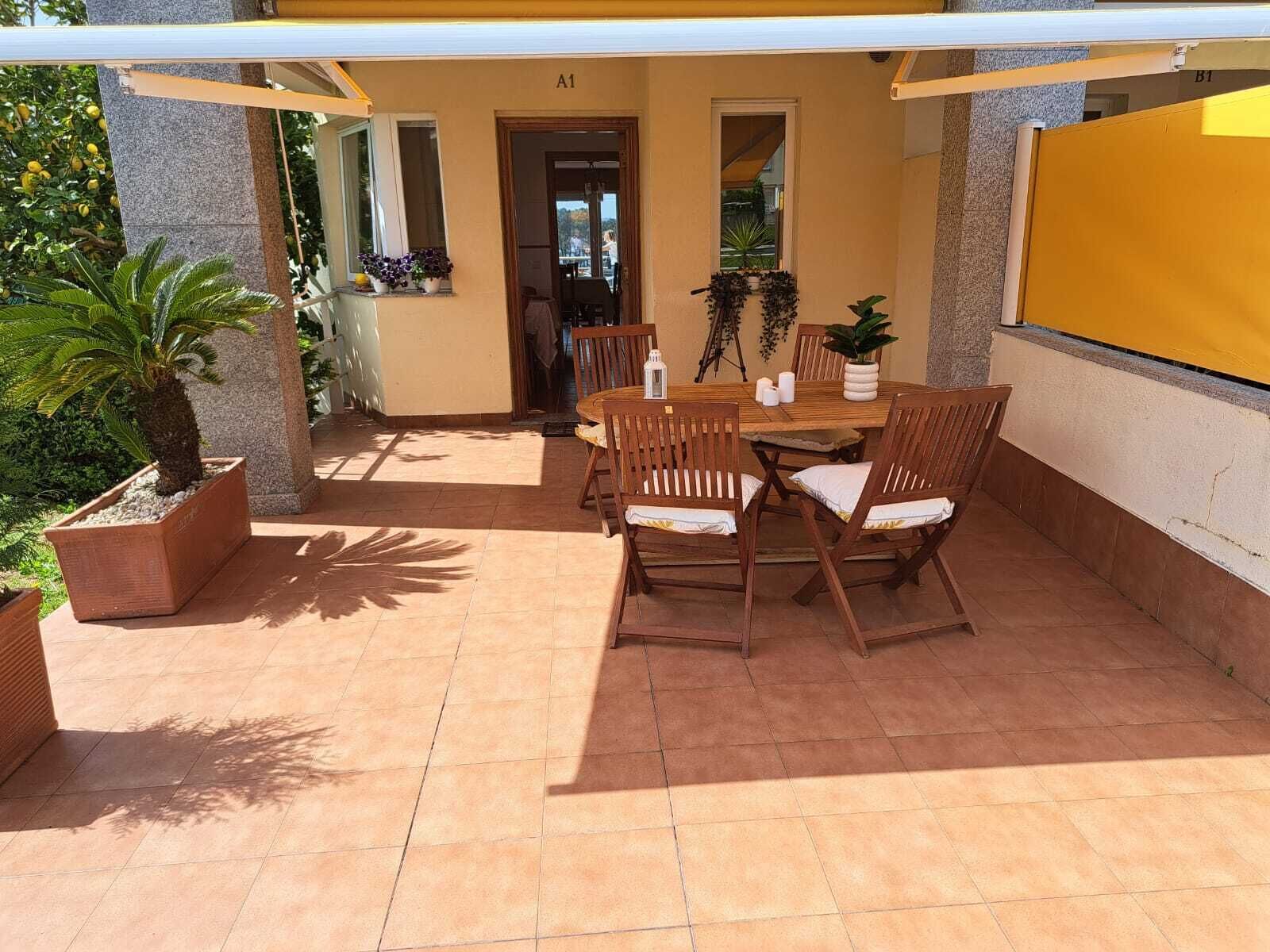 Un patio exterior con una mesa redonda de madera y seis sillas, rodeado de macetas con plantas y una pantalla amarilla.
