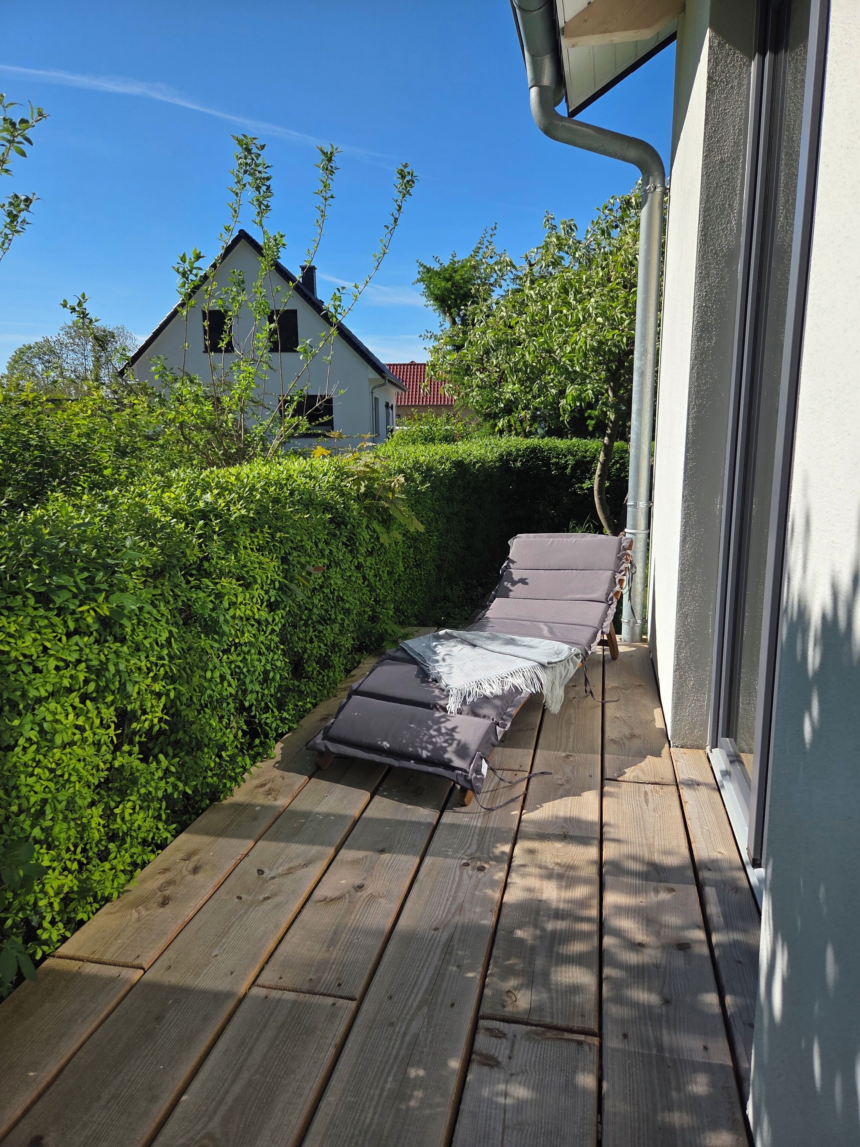 Een houten terras met een ligbed en een kleine tafel, omringd door weelderige begroeiing en een huis op de achtergrond.