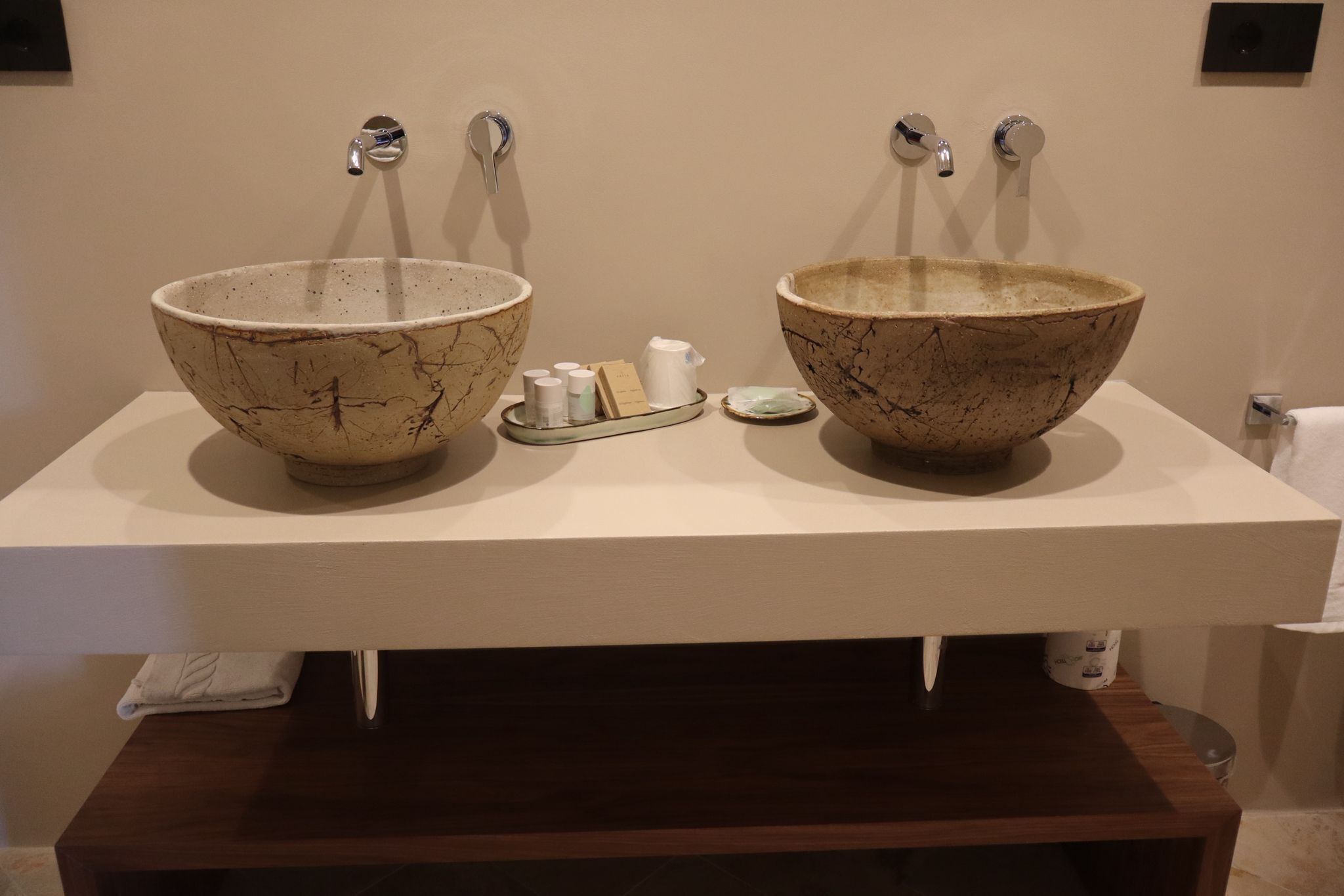 Ein Badezimmer mit zwei großen, marmorähnlichen Waschbecken, jeweils mit einem Wasserhahn. Auf einem Tablett dazwischen liegen Pflegeprodukte.