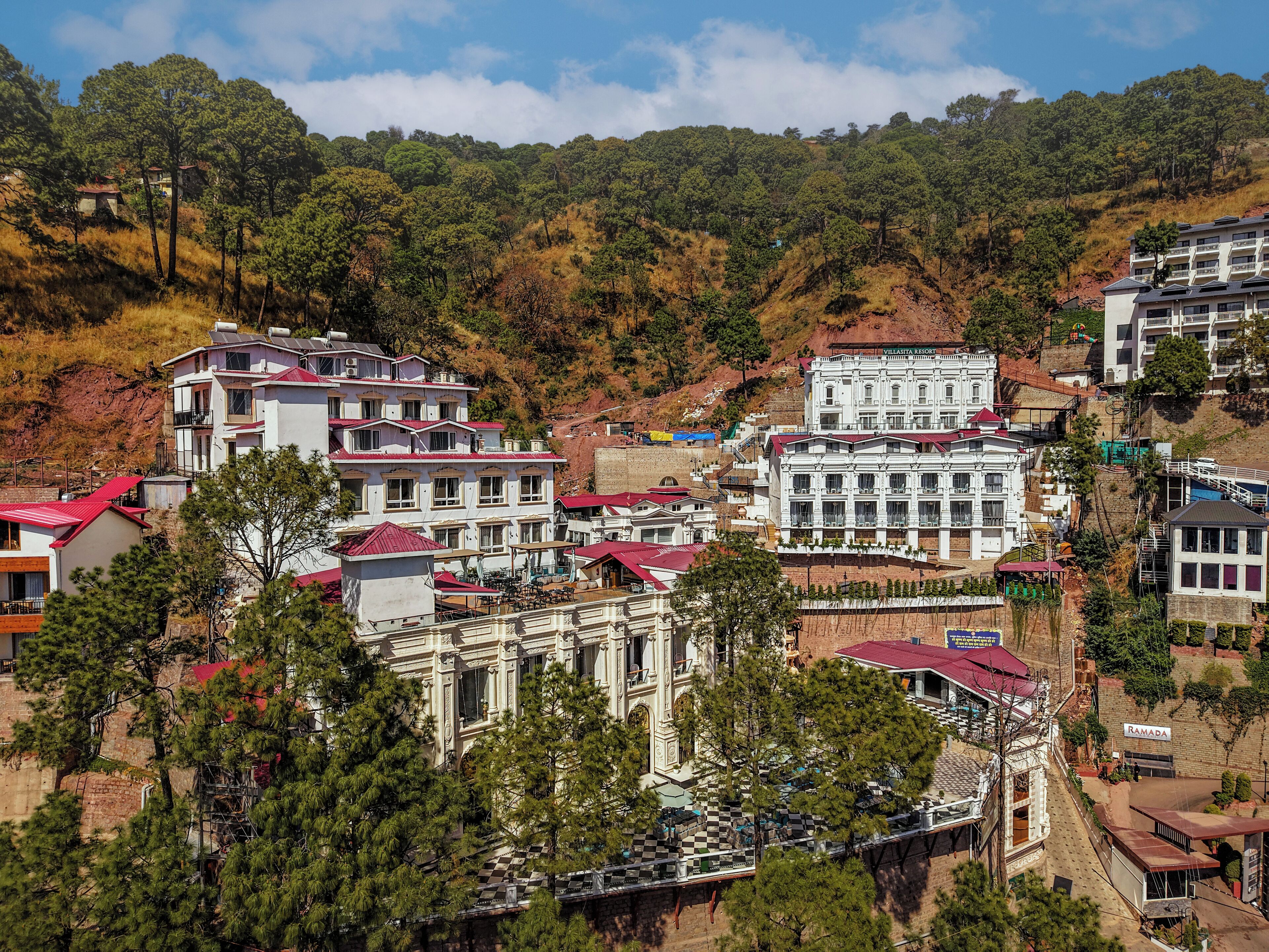 A resort with multiple buildings, including a large white building with a red roof and a smaller building with a red roof. There are trees and hills in the background.