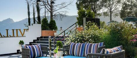 A checkered outdoor seating area with wicker furniture and cushions, a staircase leading to a higher level, and a view of mountains in the background.