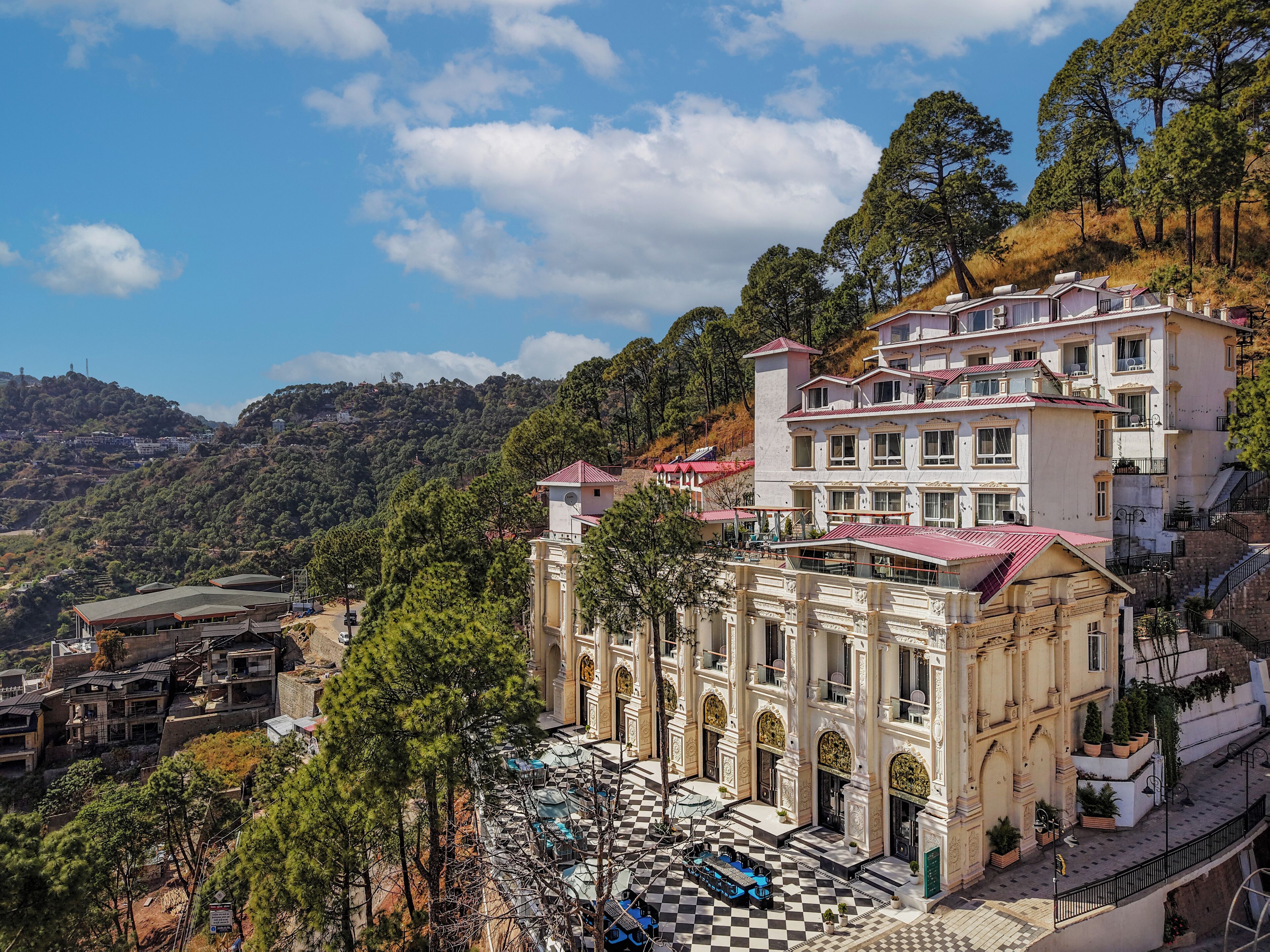 A large, historic building with a checkered courtyard and multiple balconies, surrounded by lush greenery and hills.