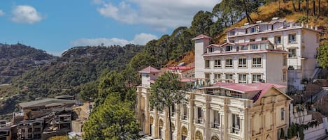 A large, historic building with a checkered courtyard and multiple balconies, surrounded by lush greenery and hills.