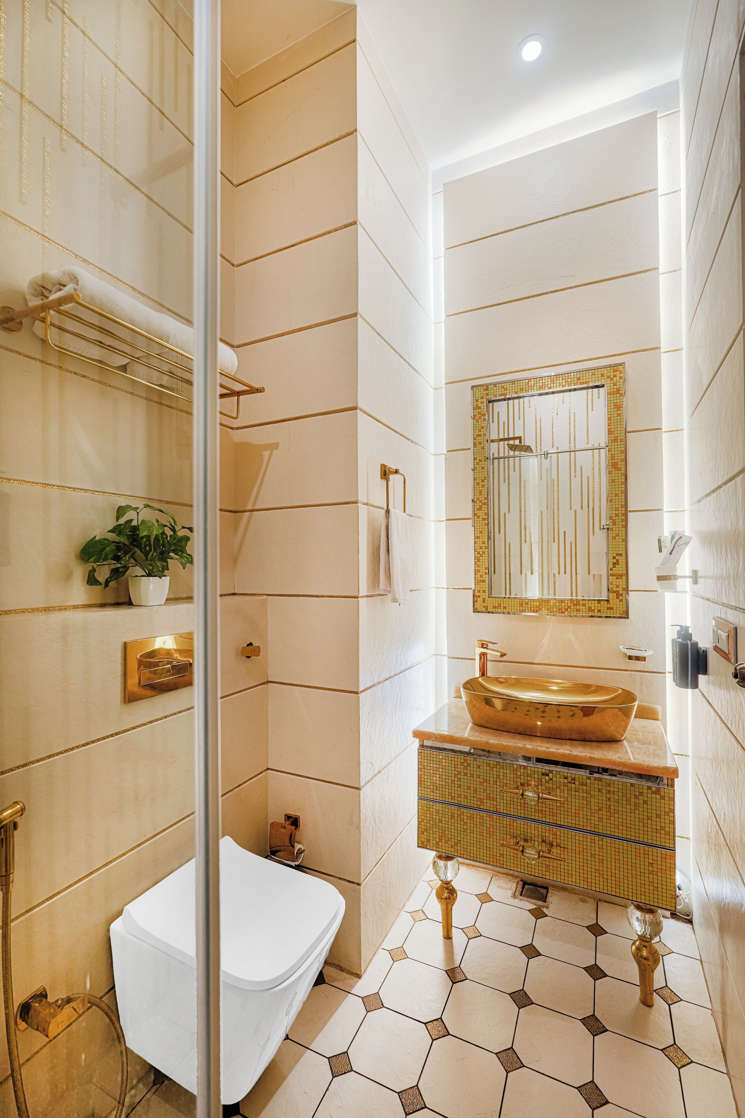 A modern bathroom with a white toilet, a glass-enclosed shower, a gold sink, and a mirror with a gold frame.