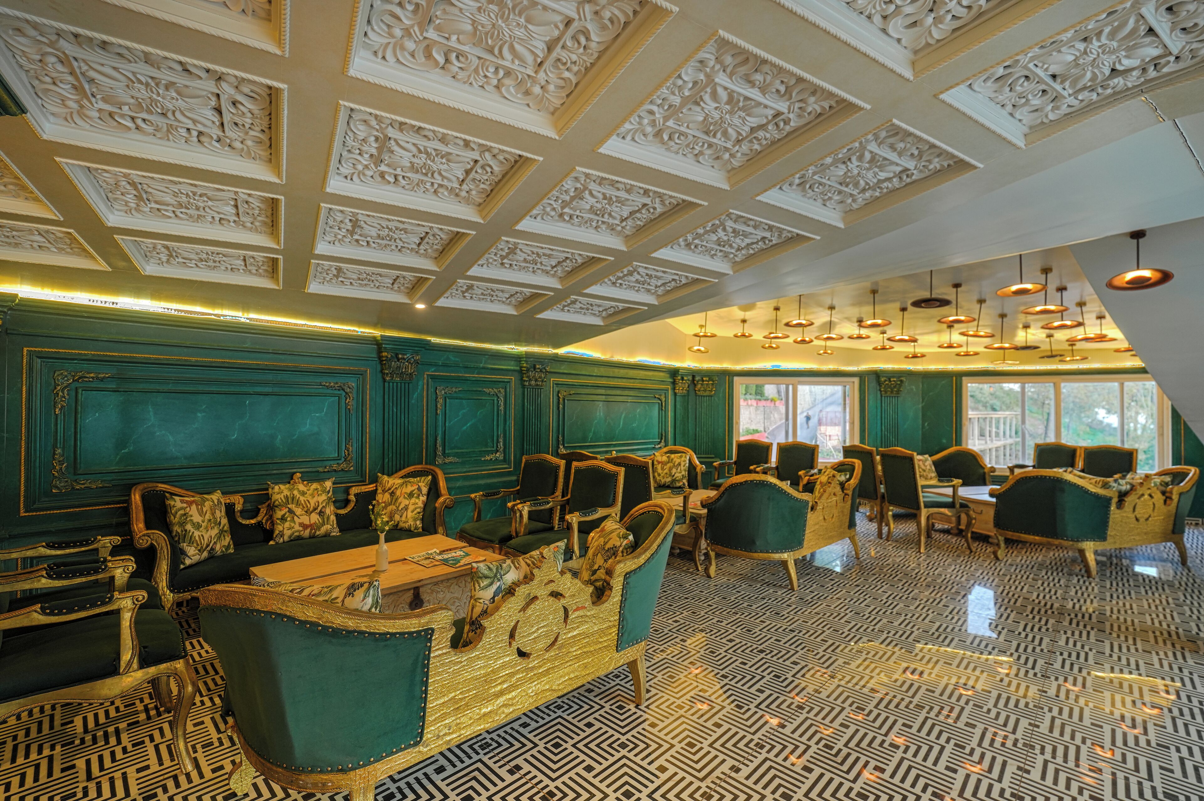 A room with ornate ceiling carvings, green upholstered furniture, and patterned flooring.