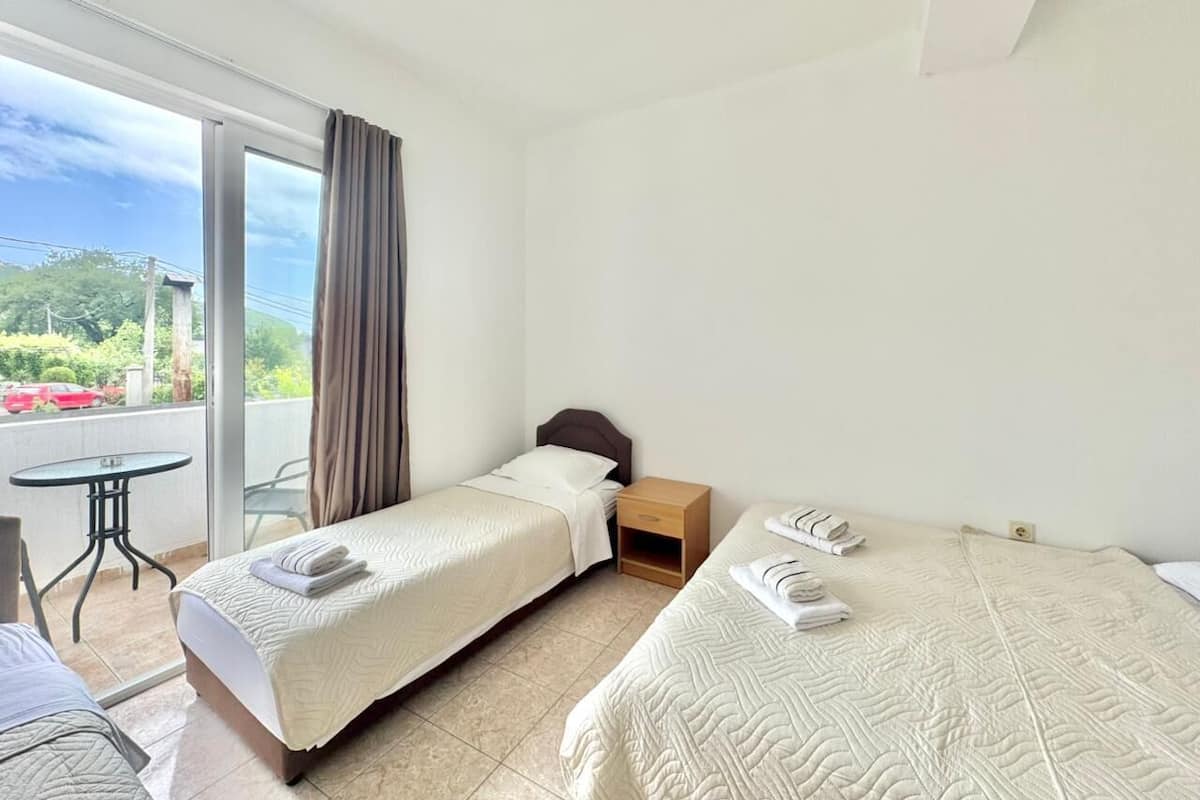 Een hotelkamer met twee bedden, een kleine tafel en een balkon met uitzicht op groen en een rode auto.