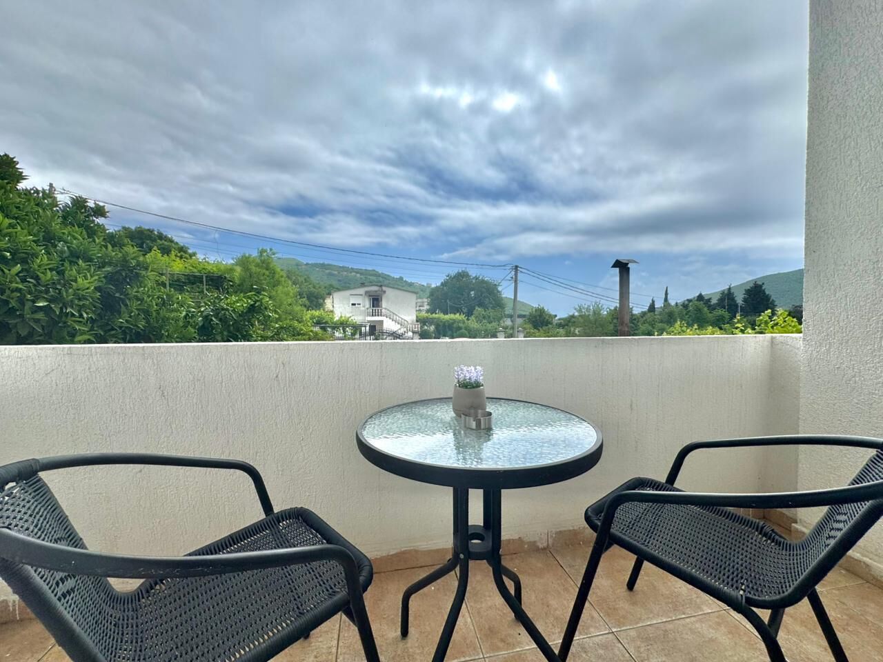 A balcony with two chairs and a small table, overlooking a residential area with houses and greenery under a cloudy sky.