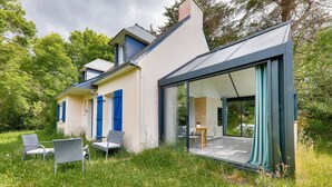 A modern house with a glass door leading to a dining area, surrounded by lush greenery.
