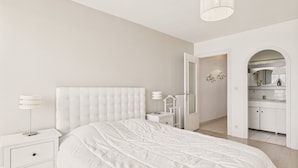 A modern bedroom with a large bed, bedside tables, a nightstand, a lamp, and an open door leading to a bathroom.