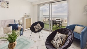 A modern living room with a sofa, chairs, a glass coffee table, and a view of the beach through large sliding doors.