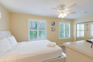 A hotel room with a bed, a desk, a ceiling fan, and two windows with blinds.