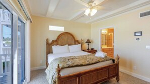 A bedroom with a wooden bed, a ceiling fan, a nightstand, a window, and a bathroom visible through an open door.