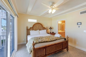 Une chambre à coucher avec un lit en bois, un ventilateur de plafond, une table de chevet, une fenêtre et une salle de bain visible à travers une porte ouverte.