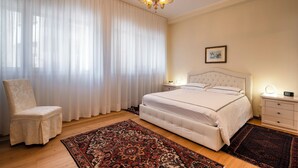 Ein Schlafzimmer mit einem großen Bett, einem Sessel, einem Nachttisch, einer Nachttischlampe, einer Chandelüster und einem Fenster mit durchsichtigen Vorhängen.