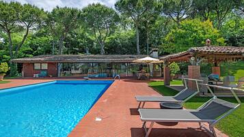 Piscina com espreguiçadeiras, alpendre coberto e um edifício de tijolos com telhado em cerâmica.