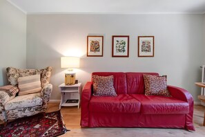 A living room with a red sofa, floral armchair, and a patterned rug. There are three framed artworks on the wall and a lamp on a side table.