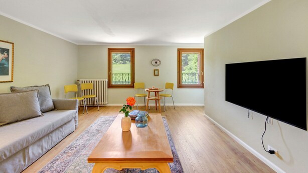 Ein Wohnzimmer mit Sofa, Couchtisch, zwei Sesseln, Fernseher und Fenster mit Blick.