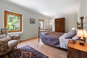 A bedroom with a bed, a chair, a bedside table with a lamp, a radiator, and a window with a view of greenery.