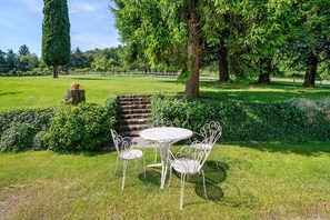 Un jardín con una mesa y sillas de metal blanco, rodeado de abundante vegetación y árboles.