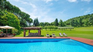 Ein Schwimmbecken mit Liegestühlen und einer Pergola, umgeben von einer Rasenfläche und Bäumen.