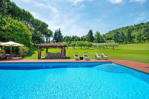 Una piscina con sillones de descanso y una pérgola, rodeada de césped y árboles.