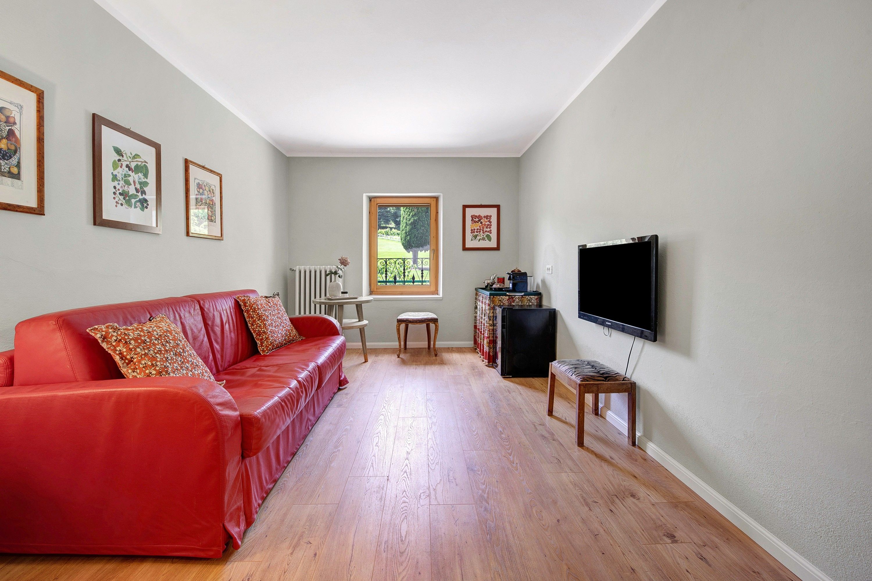 Ein Wohnzimmer mit einem roten Sofa, einem Fernseher, gerahmten Bildern an der Wand und einem Fenster mit Blick auf Grünflächen.