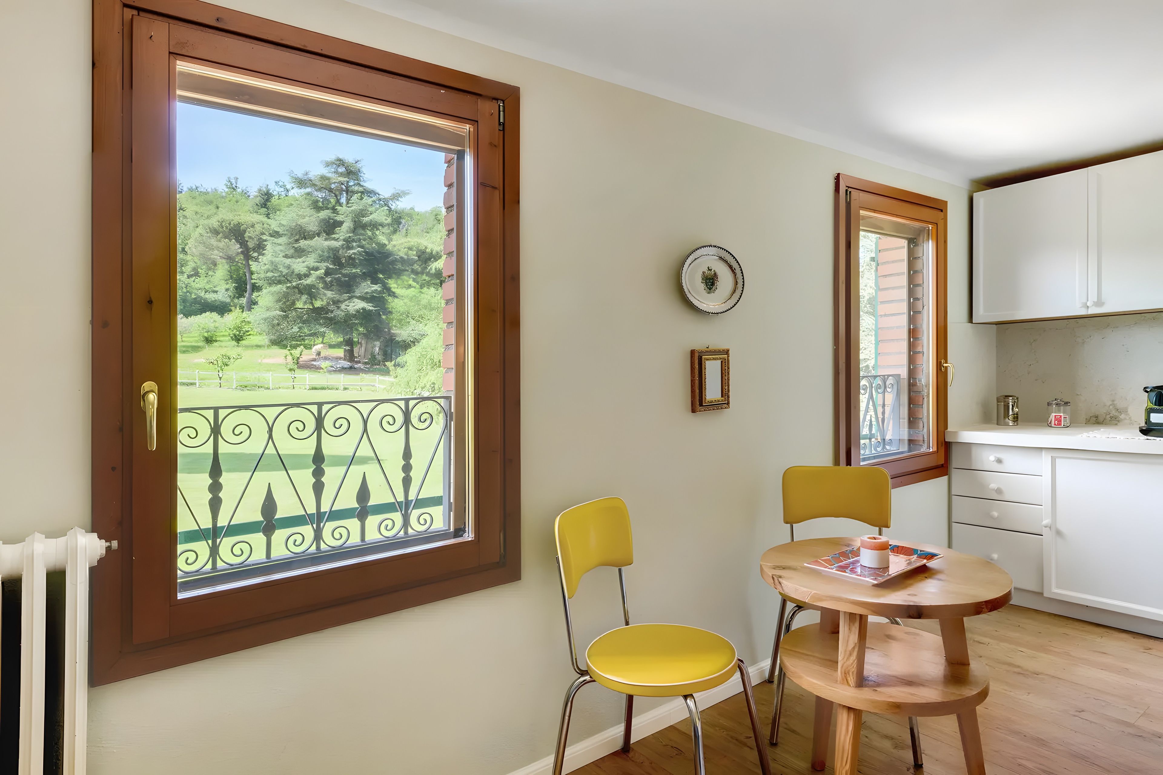 A room with a dining table, two chairs, a window with a view of a garden, and a kitchen area with cabinets and a countertop.
