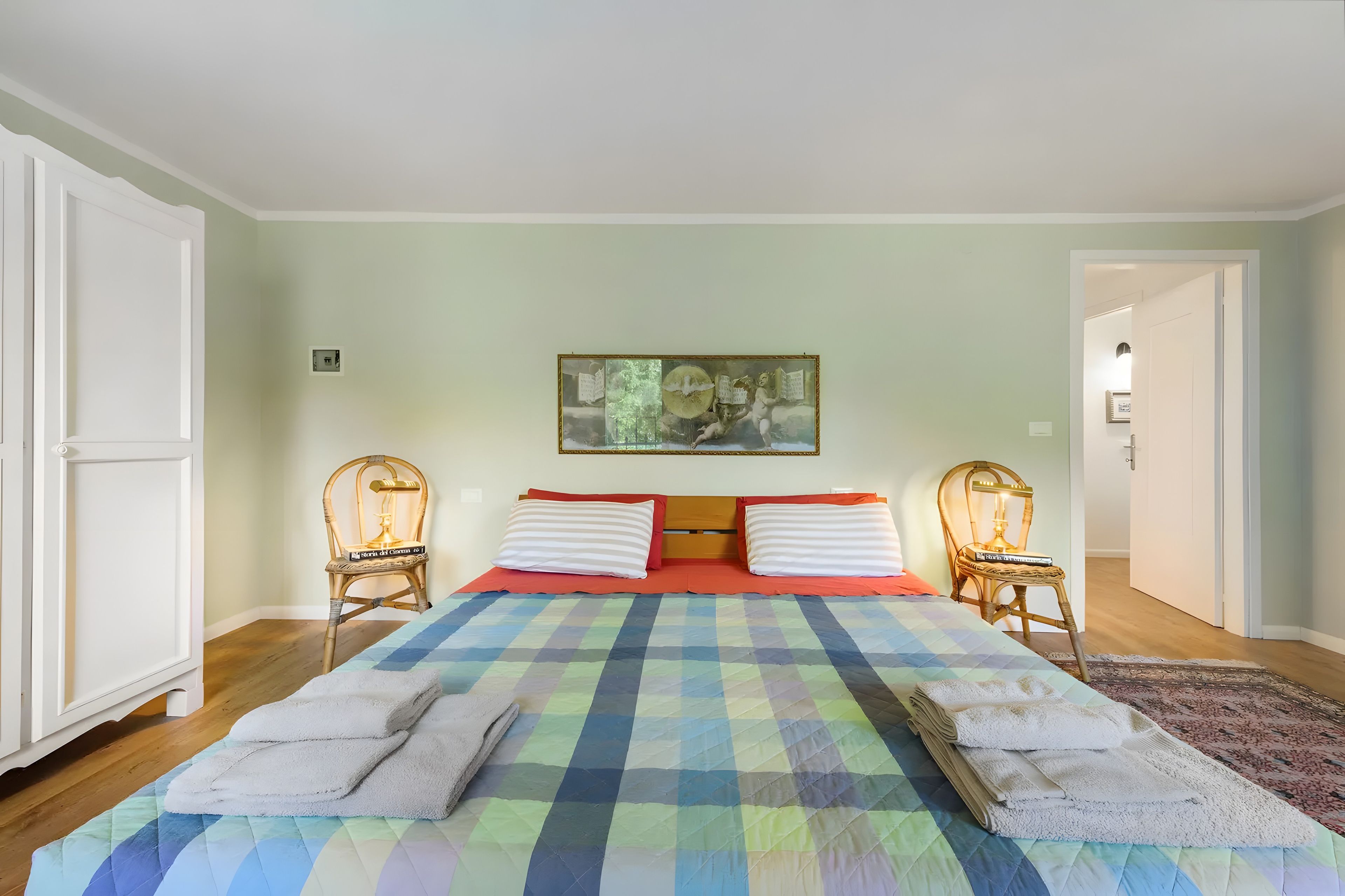 Un dormitorio con una cama grande, dos sillas, un cuadro en la pared y una puerta a la derecha.