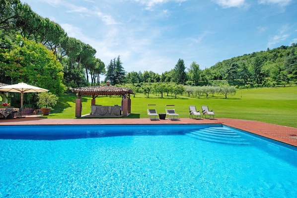 A swimming pool with lounge chairs and a pergola, surrounded by a grassy area and trees.