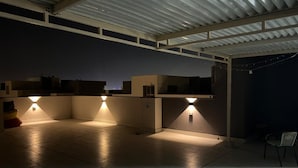 Un espacio al aire libre techado, con piso de baldosas, techo de metal y tres luces fijadas a la pared.