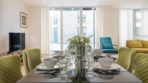 A dining area with a table set for four, a blue armchair, a yellow sofa, and a large window with a view of a brick building. - Austin David Royal Wharf  (London)