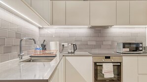 A modern kitchen with white cabinetry, a stainless steel sink, a kettle, a microwave, and a built-in oven. - Austin David Royal Wharf  (London)