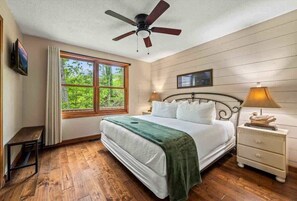 Quarto com cama, ventilador de teto, janela com cortinas, mesa de cabeceira e criado-mudo.