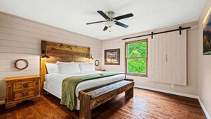 A bedroom with a wooden bed, a bench, a nightstand, a ceiling fan, and a window with a view of greenery.