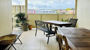 Un balcon abrité, avec une table et des chaises en bois, un pot de fleurs et une vue sur un bâtiment portant l’inscription « Harvey Norman ».