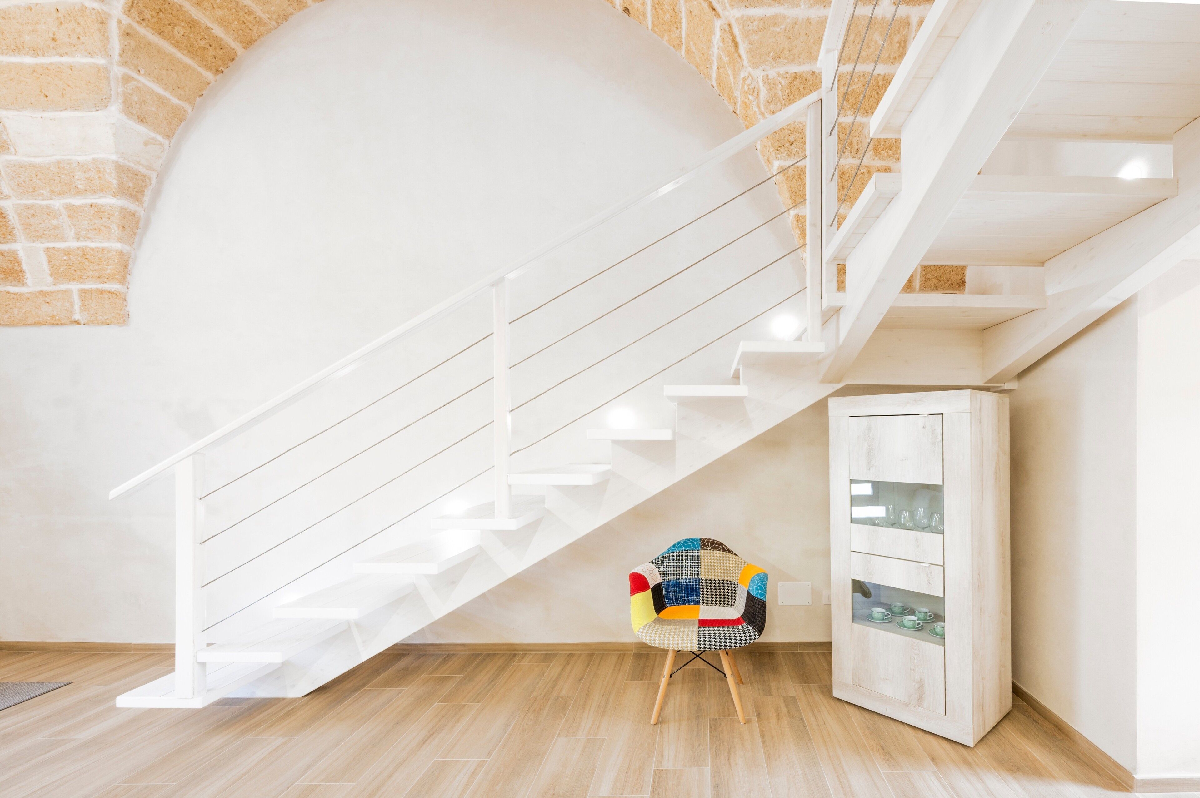 A modern interior with a white staircase, a colorful chair, and a cabinet with glass doors.