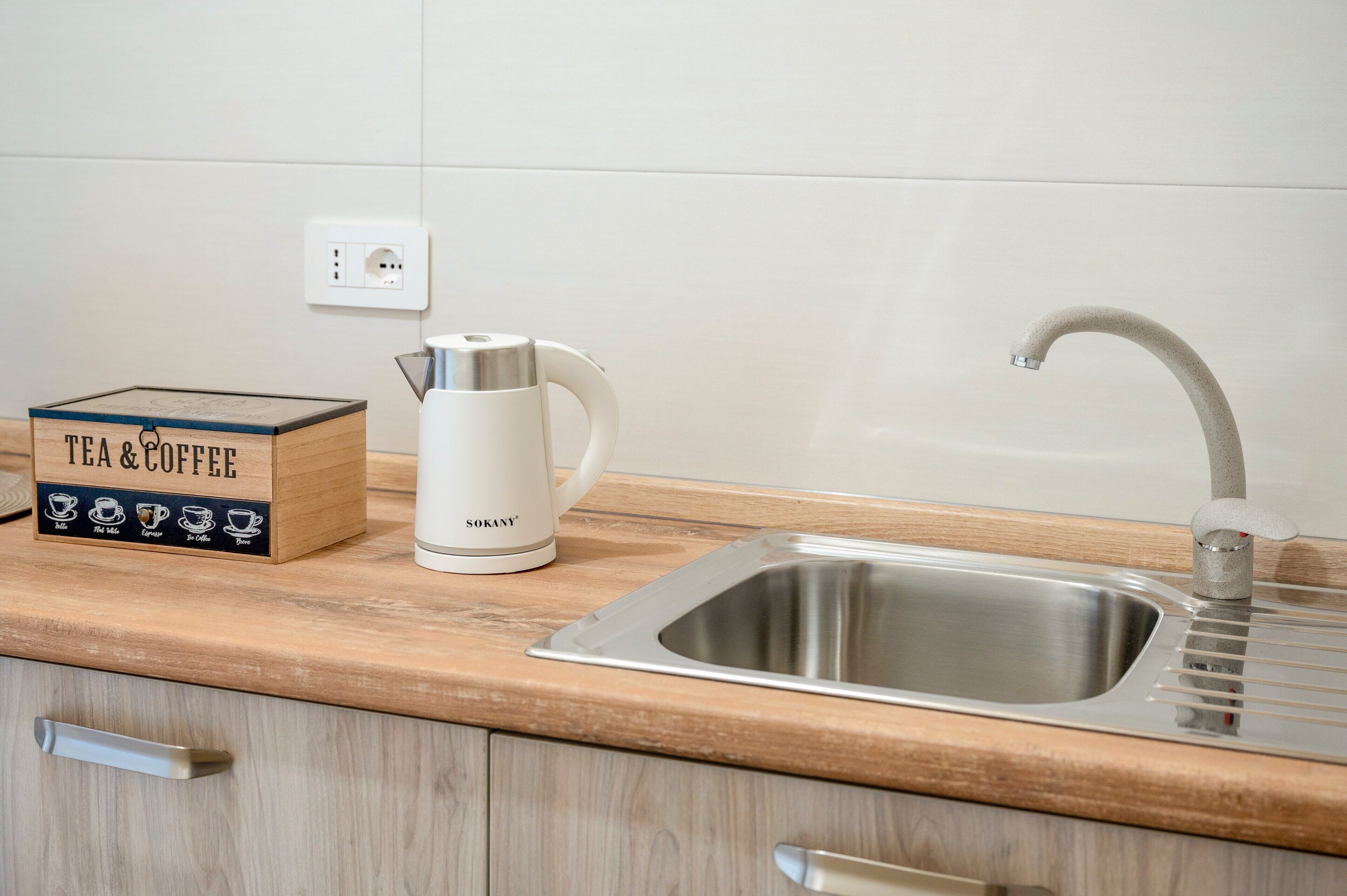 Una cocina con un lavamanos de acero inoxidable, una tetera blanca y una caja para té y café en la encimera.