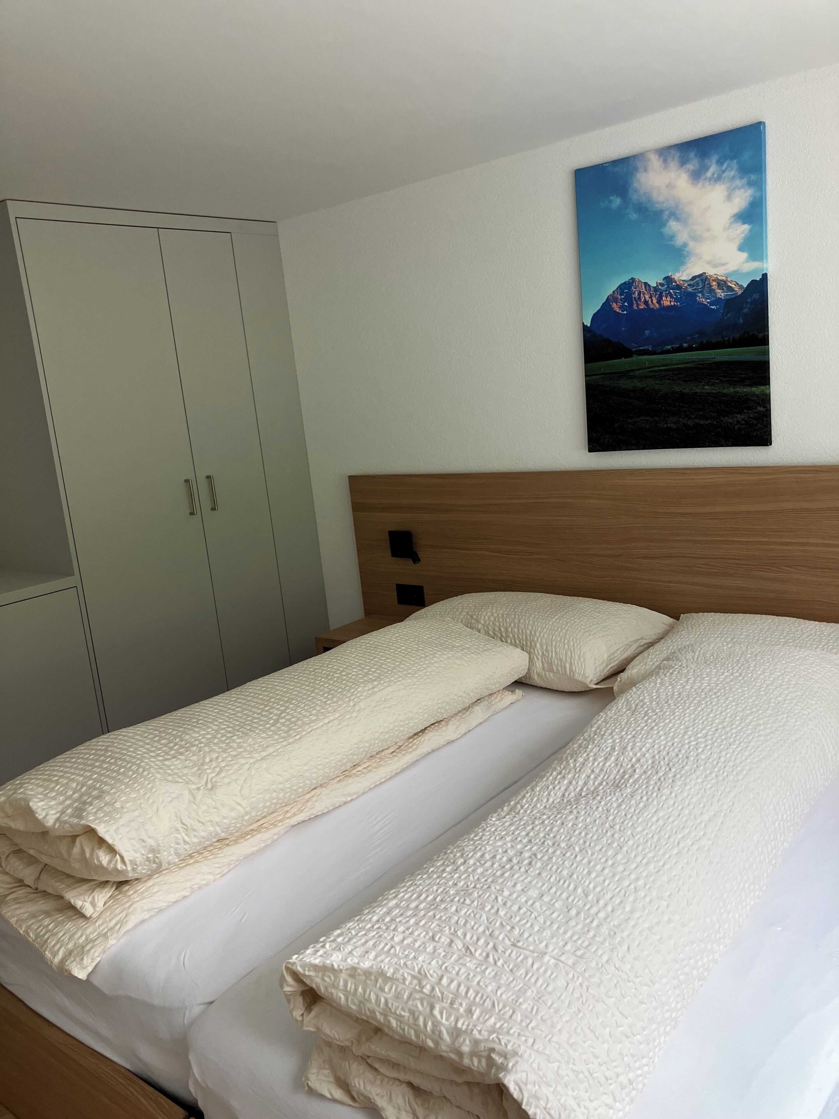 A neatly made bed with a textured white duvet, a wooden headboard, and a mountain landscape painting above it.