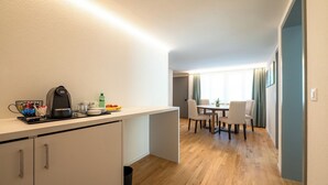 Quarto de hotel compacto com cozinha americana, área de refeições com mesa e cadeiras, e vista para o exterior pela janela.