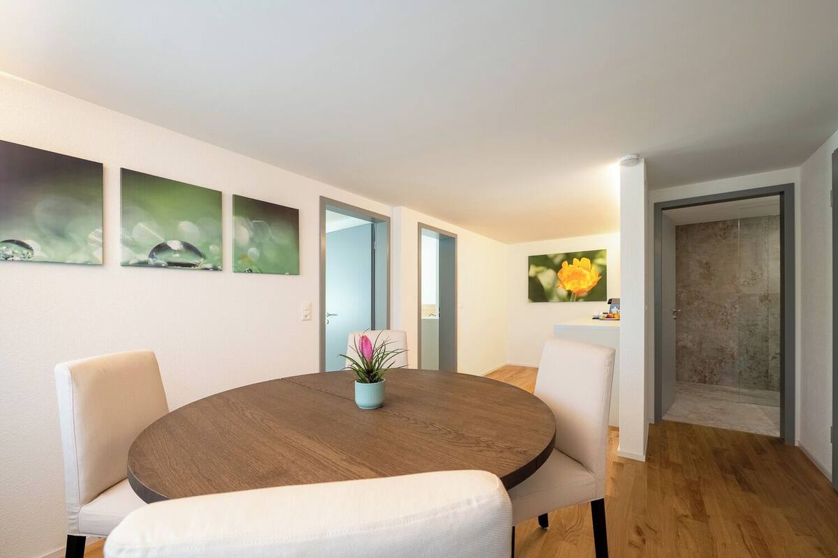 Een moderne eethoek met een ronde houten tafel, witte beklede stoelen en een potplant.