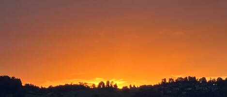A sunset over a hilly landscape with a silhouette of trees and buildings.