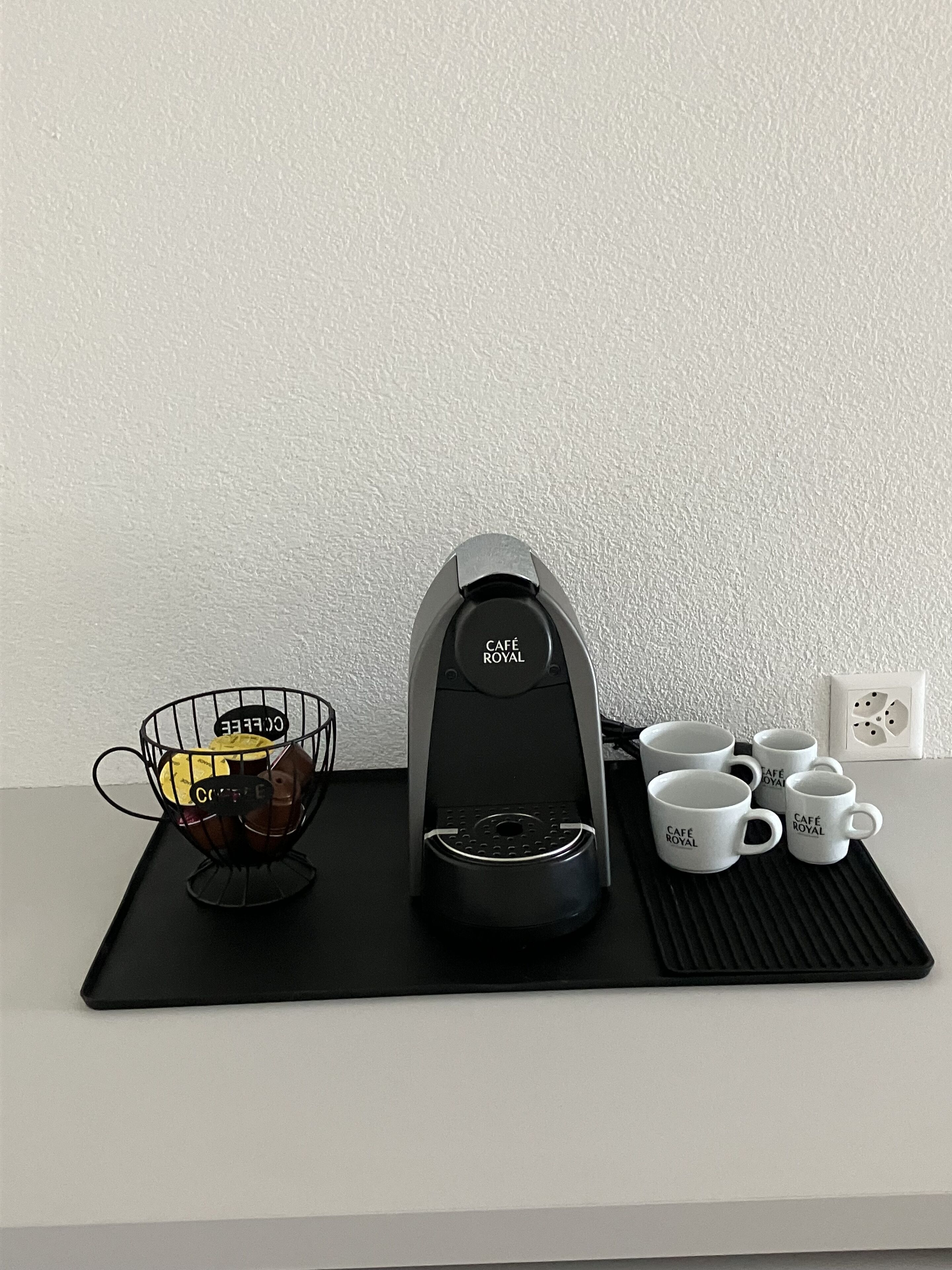 A coffee maker, a wire basket with coffee pods, and four white mugs on a black tray.