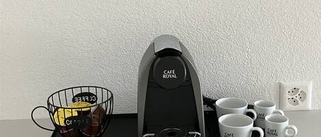 A coffee maker, a wire basket with coffee pods, and four white mugs on a black tray.