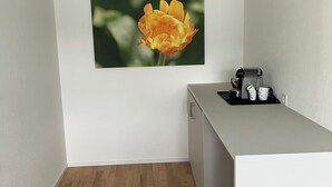 Quarto pequeno e limpo, com piso de madeira, uma escrivaninha branca com máquina de café e um quadro de uma flor na parede.