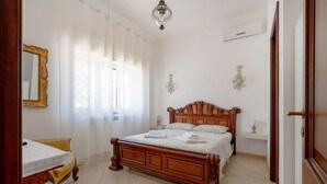 A bedroom with a wooden bed, a bedside table, a chair, a window with curtains, and a chandelier.