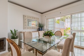 A dining area with a glass table, beige chairs, a potted plant, and a decorative mirror.