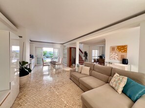A spacious living area with a beige sofa, a glass coffee table, and a view of a dining area with a staircase. - Villa 'Casa Do Rei - Charneca' with Private Pool, Private Terrace and Wi-Fi (Charneca de Caparica)