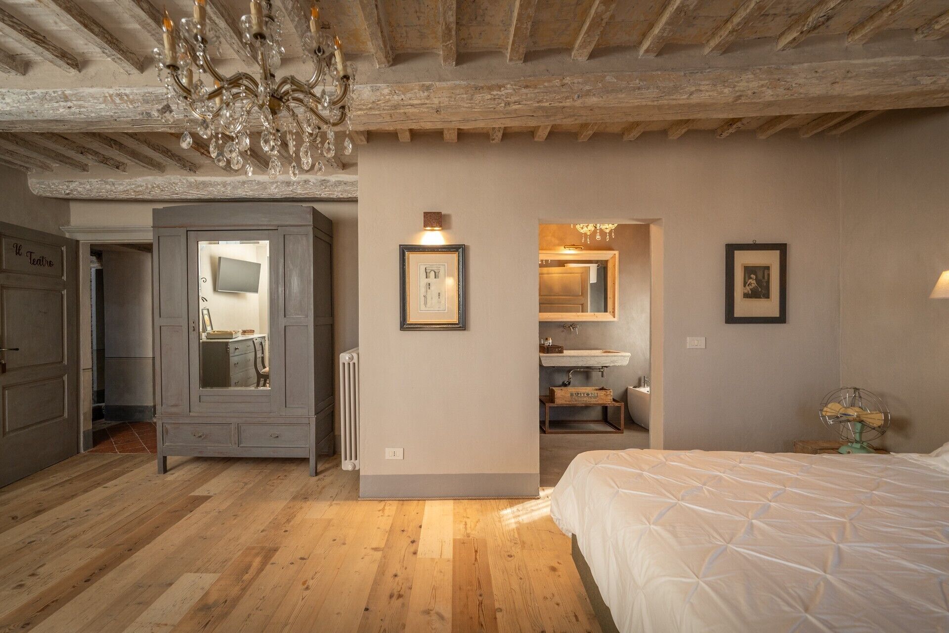 Un dormitorio con suelo de madera, una cama, una araña y un aparador; se ve un baño a través de una puerta abierta.
